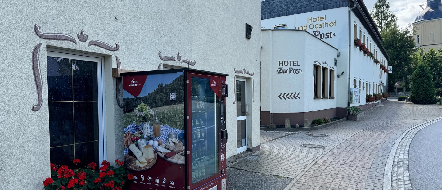 Automat na potraviny v hotelu "Zur Post" Před hotelem s nápisem "Hotel Zur Post" stojí červený automat a vedle něj kvetou červené muškáty.
