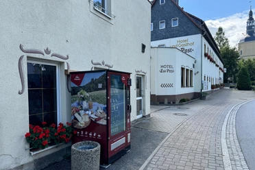 Proviantomat Standort am Hotel "Zur Post" Ein roter Verkaufsautomat steht vor einem Hotel mit der Aufschrift "Hotel Zur Post", daneben blühende rote Geranien.