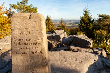 BN6_6c_Velmerstot Route © Tourismus NRW e.V. (14).jpg Stein mit Inschrift und Blick auf hügelige Landschaft unter blauem Himmel, umgeben von Bäumen.