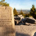 BN6_6c_Velmerstot Route © Tourismus NRW e.V. (14).jpg Stein mit Inschrift und Blick auf hügelige Landschaft unter blauem Himmel, umgeben von Bäumen.