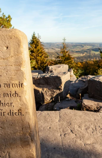 BN6_6c_Velmerstot Route © Tourismus NRW e.V. (14).jpg Stein mit Inschrift und Blick auf hügelige Landschaft unter blauem Himmel, umgeben von Bäumen.