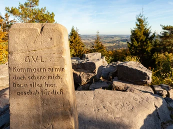 BN6_6c_Velmerstot Route © Tourismus NRW e.V. (14).jpg Stein mit Inschrift und Blick auf hügelige Landschaft unter blauem Himmel, umgeben von Bäumen.