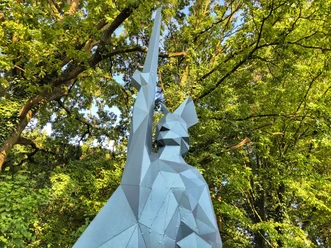 Kantiger Hermann Skulptur eines stilisierten, kantigen Hermann mit hoch erhobenem Schwert vor dichtem Laubwerk.