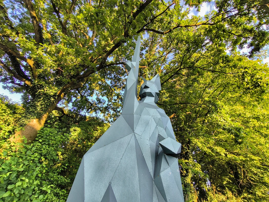 Kantiger Hermann Skulptur des kantigen Hermann aus Metall vor üppig grünen Bäumen unter strahlend blauem Himmel.