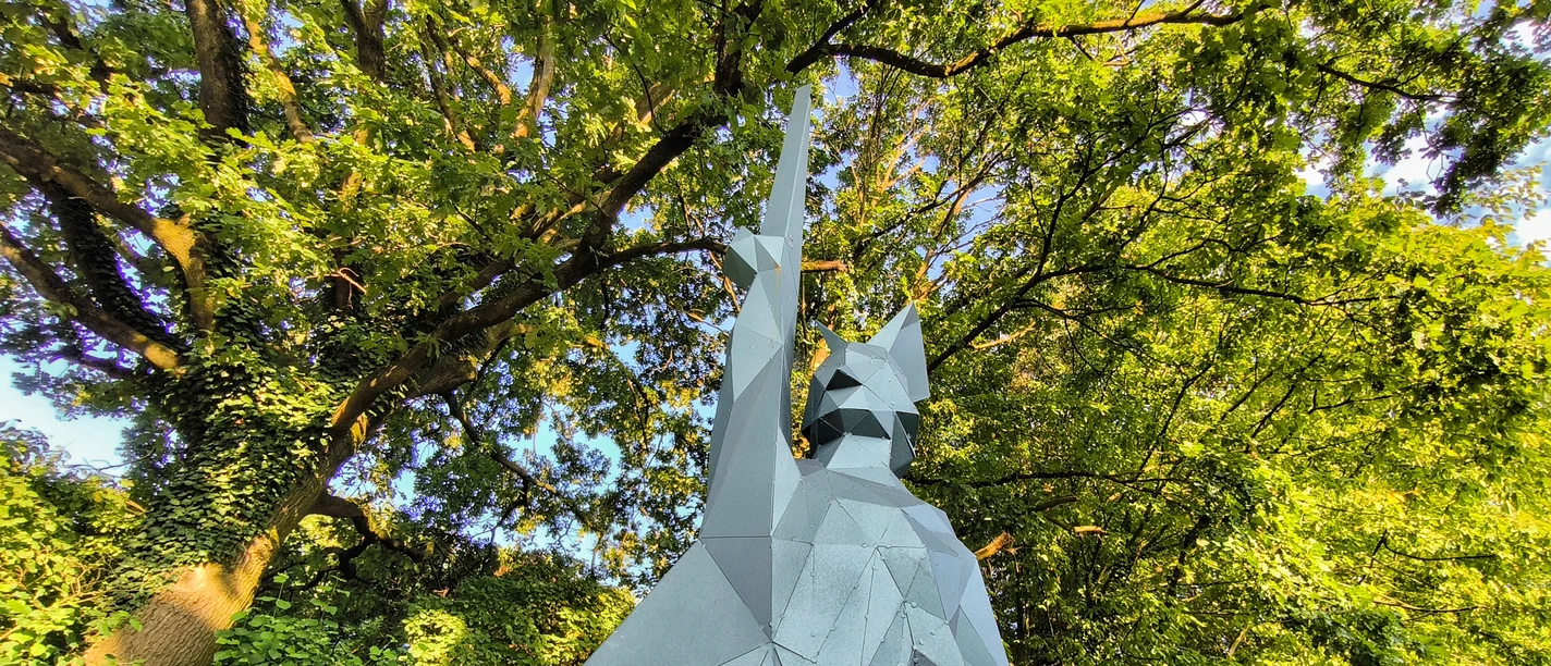 Kantiger Hermann Skulptur des kantigen Hermann aus Metall vor üppig grünen Bäumen unter strahlend blauem Himmel.