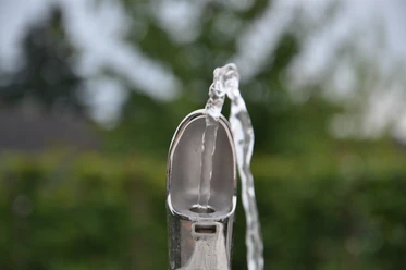 Trinkwasserspender_Greffen 009.jpg Ein silberner Trinkbrunnen sprudelt klares Wasser, umgeben von unscharfem Grün im Hintergrund.