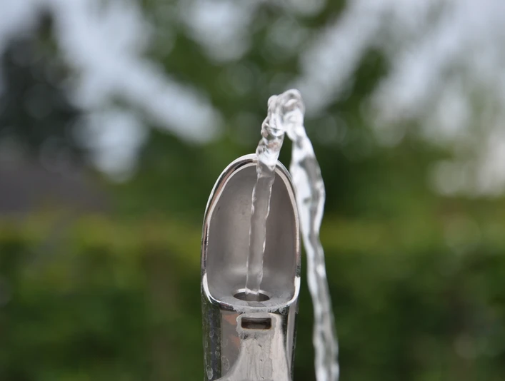Trinkwasserspender_Greffen 009.jpg Ein silberner Trinkbrunnen sprudelt klares Wasser, umgeben von unscharfem Grün im Hintergrund.