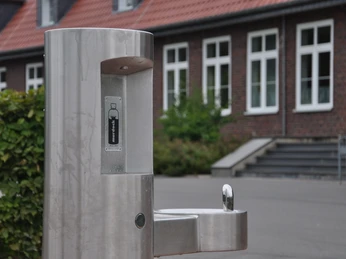 Trinkwasserspender_Greffen 017.jpg Öffentlicher Trinkbrunnen aus Edelstahl vor einem Backsteingebäude mit weißen Fensterrahmen.