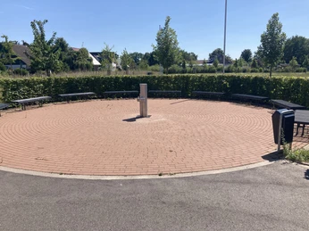 IMG_6031.JPG Öffentlicher runder Platz mit Ziegelpflaster, zentraler Wasserquelle und umgebenden Sitzbänken.