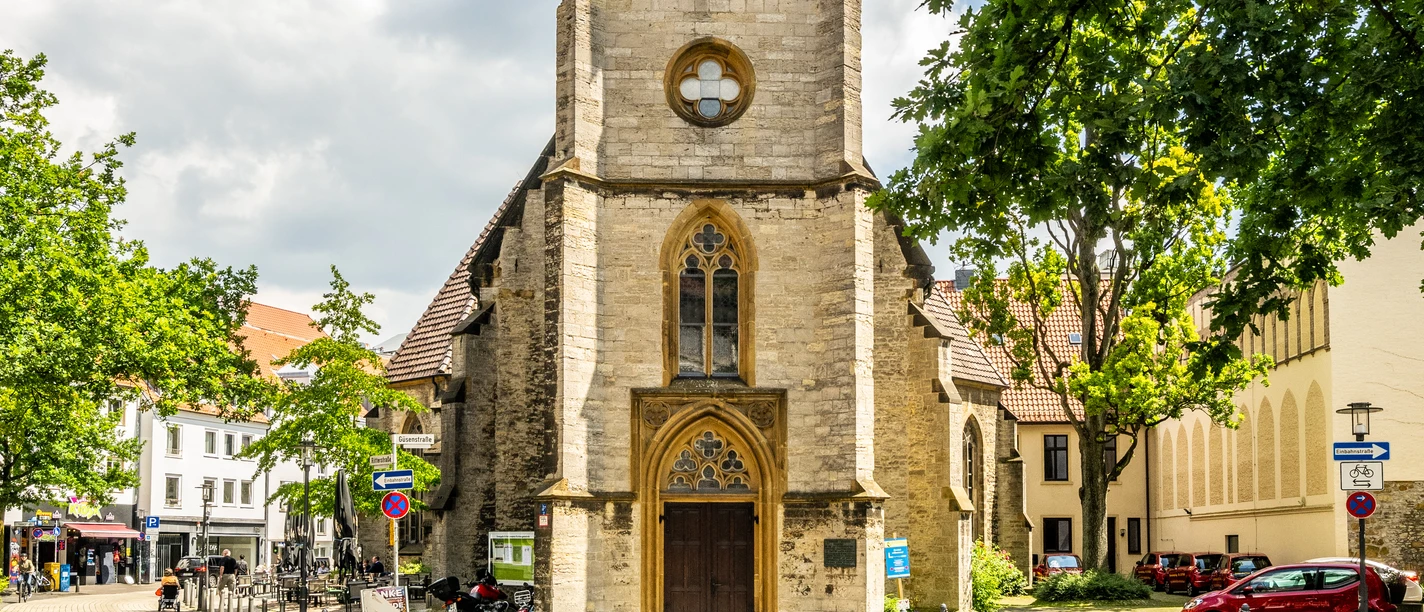Historische Kirche mit hohem Turm, umgeben von Bäumen und blauem Himmel in urbanem Umfeld.