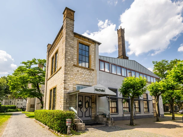 Historisches Fabrikgebäude mit modernem Anbau, umgeben von Grünflächen und blauem Himmel im Hintergrund.