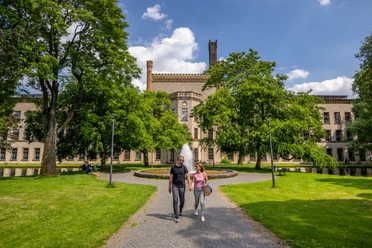 Ravensberger Park Bielefeld Ein Paar spaziert auf einem gepflasterten Weg durch eine grüne Allee vor einem historischen Gebäude.