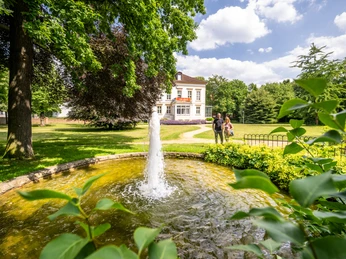 Historische Villa mit Springbrunnen in sonnigem Garten, umgeben von grünen Bäumen und Spaziergängern.