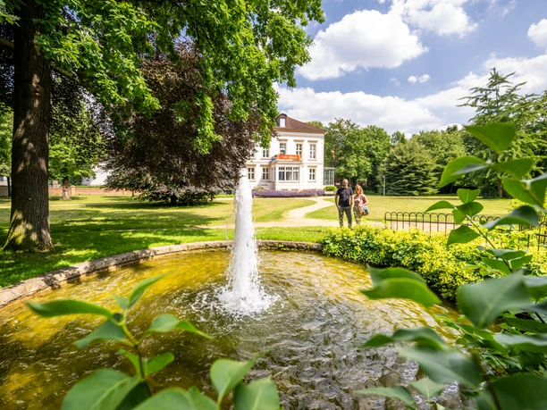 Historische Villa mit Springbrunnen in sonnigem Garten, umgeben von grünen Bäumen und Spaziergängern.
