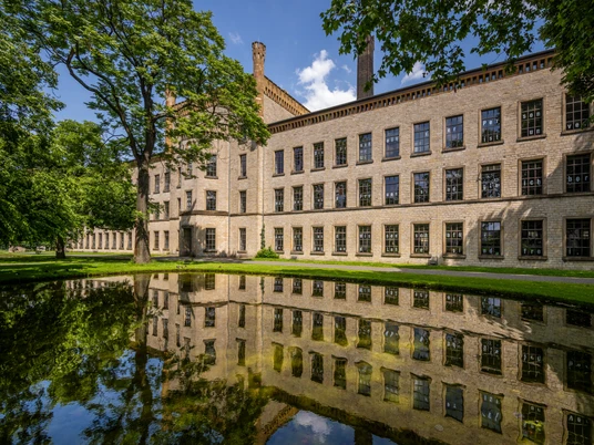 Ein historisches Backsteingebäude spiegelt sich malerisch in einem ruhigen Teich, umgeben von Grün.