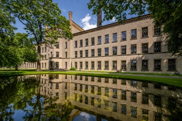 Ravensberger Spinnerei Bielefeld Ein historisches Backsteingebäude spiegelt sich malerisch in einem ruhigen Teich, umgeben von Grün.