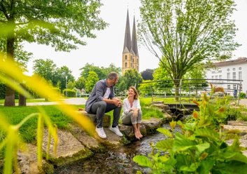 Park der Menschenrechte Bielefeld Ein Paar sitzt an einem kleinen Bach in einem Park, mit einer Kirche im Hintergrund.