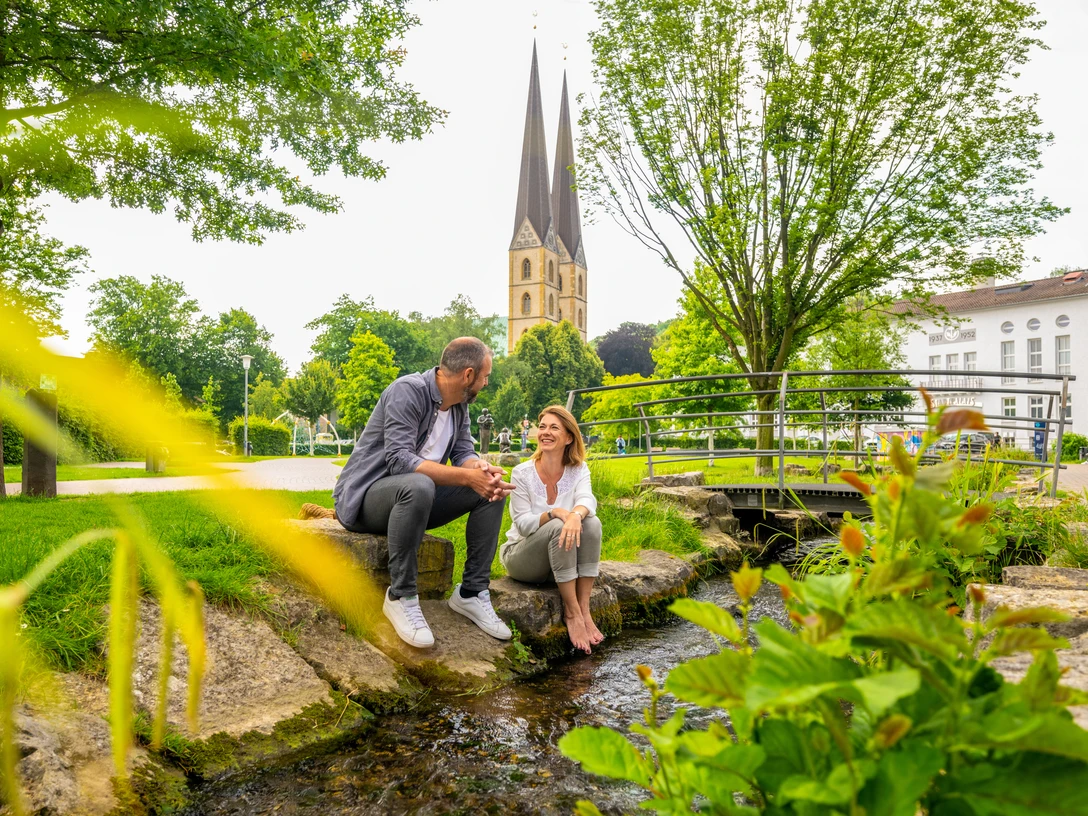Park der Menschenrechte Bielefeld Ein Paar sitzt an einem kleinen Bach in einem Park, mit einer Kirche im Hintergrund.