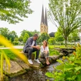Park der Menschenrechte Bielefeld Ein Paar sitzt an einem kleinen Bach in einem Park, mit einer Kirche im Hintergrund.