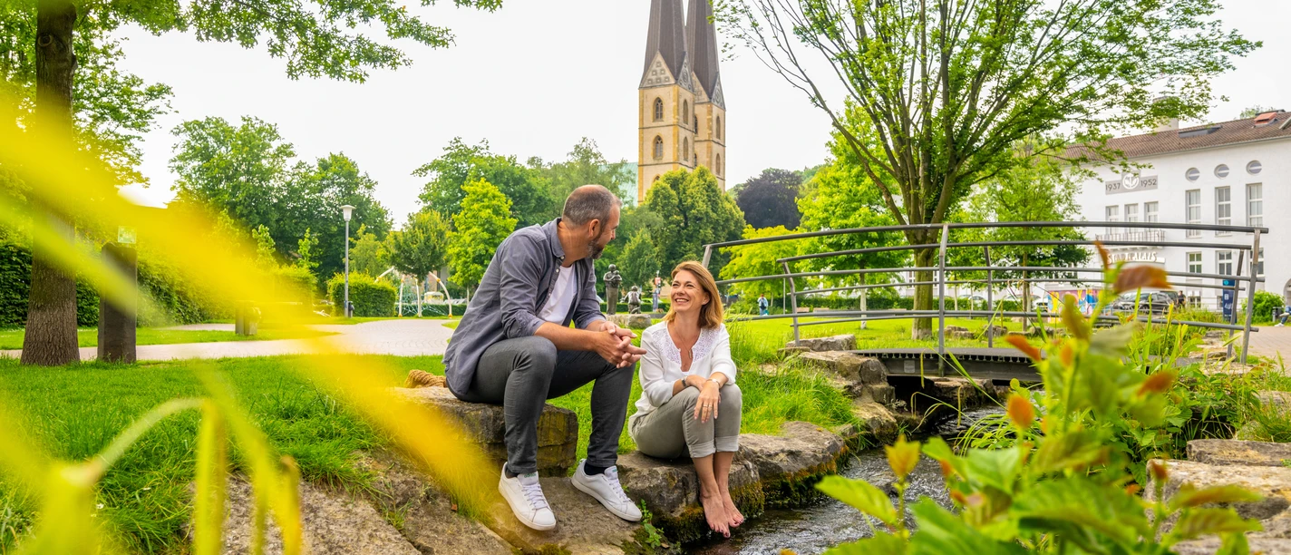 Park der Menschenrechte Bielefeld Ein Paar sitzt an einem kleinen Bach in einem Park, mit einer Kirche im Hintergrund.