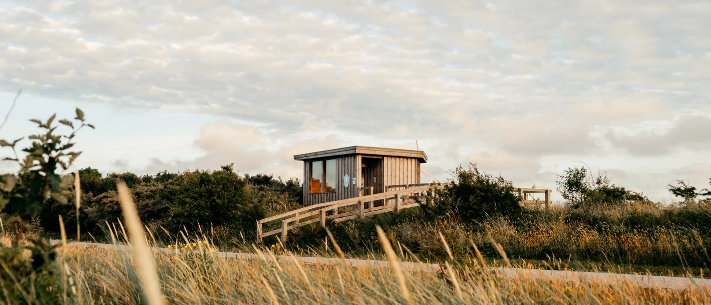 Kiekhütte.jpg Holzhütte mit Glasfront liegt in sanfter Dünenlandschaft unter einem blauen Himmel mit Wolken.