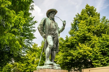 Bronzestatue eines Mannes in traditioneller Tracht mit Pfeife, umgeben von grünen Bäumen.