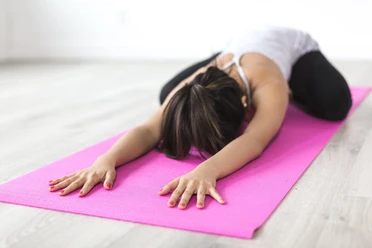 Yoga Person praktiziert Yoga-Übung "Kindshaltung" auf pinkfarbener Matte in hellem Raum.