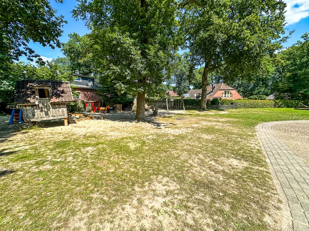 Kindergarten Oldendorf-Luhe (1).jpg Spielplatz im Freien mit Holzspielhaus, Sandfläche und schattenspendenden Bäumen.