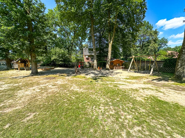 Kindergarten Oldendorf-Luhe (2).jpg Großer Spielplatz mit alten Bäumen, Holzspielhäuschen und Schaukeln auf einer Rasenfläche.