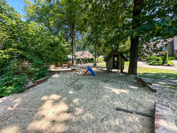 Großer Sandkasten mit Spielhaus und Rutsche im grünen Außenbereich des Kindergartens.