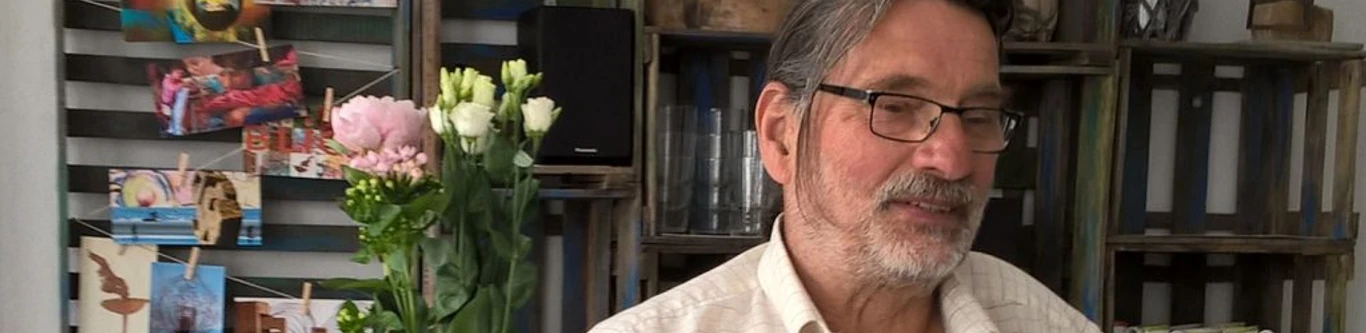 Ein älterer Mann mit Brille in einem Wohnzimmer, das mit Blumen und Bildern dekoriert ist.