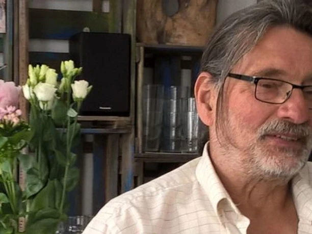 artminden.jpg Ein älterer Mann mit Brille in einem Wohnzimmer, das mit Blumen und Bildern dekoriert ist.