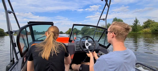 Bootsvermietung NienBoot Zwei Personen fahren ein Motorboot auf einem ruhigen Fluss, umgeben von Bäumen und blauem Himmel.