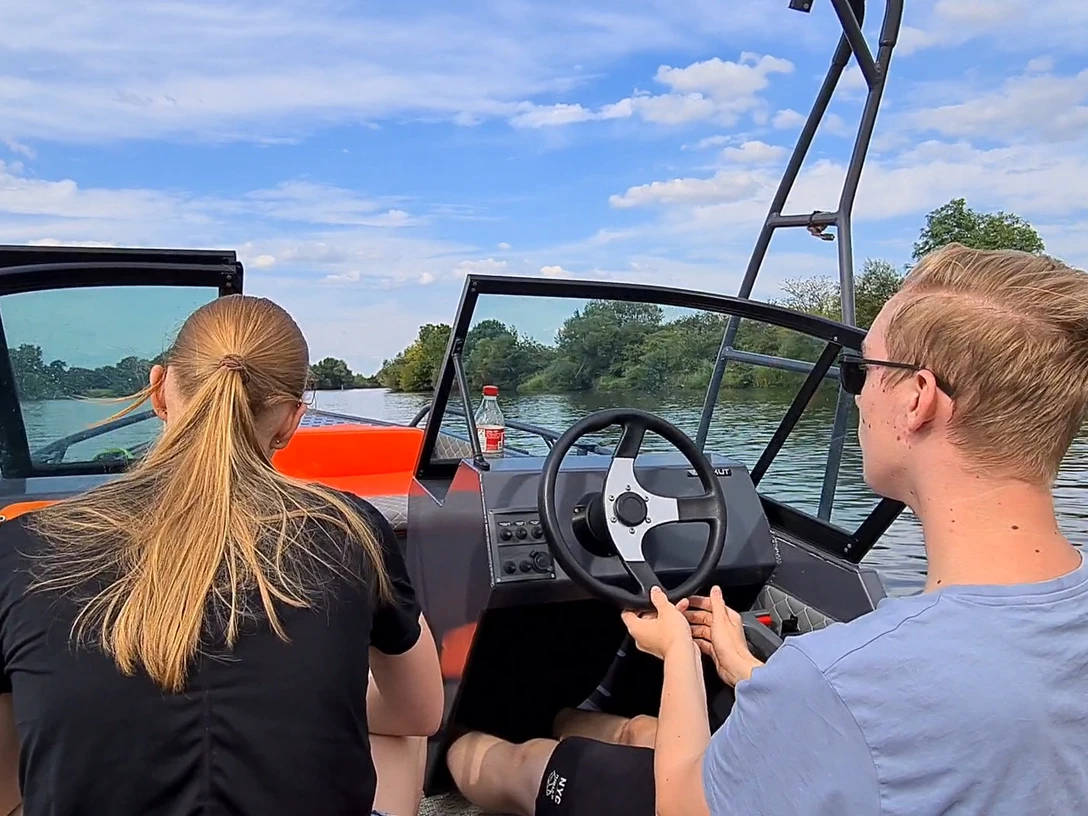 Bootsvermietung NienBoot Zwei Personen fahren ein Motorboot auf einem ruhigen Fluss, umgeben von Bäumen und blauem Himmel.