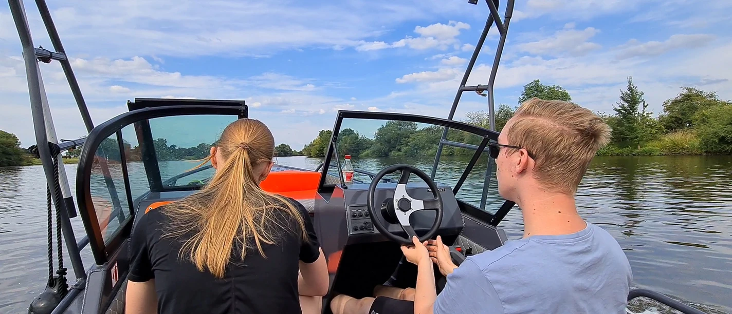 Bootsvermietung NienBoot Zwei Personen fahren ein Motorboot auf einem ruhigen Fluss, umgeben von Bäumen und blauem Himmel.