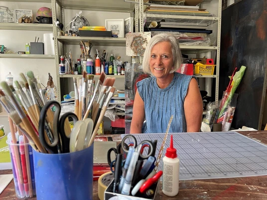MANUART.jpg Künstlerin mit grauem Haar sitzt lächelnd in ihrem Atelier, umgeben von Kunstmaterialien und Werkzeugen.