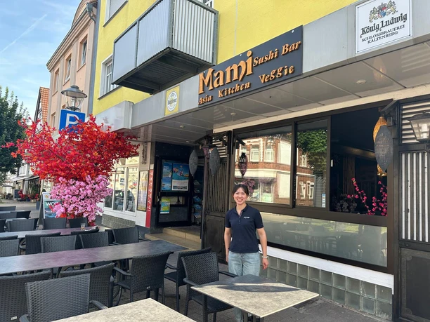 Eine Frau lächelt vor der Eingangstür eines asiatischen Restaurants mit Außenbestuhlung.