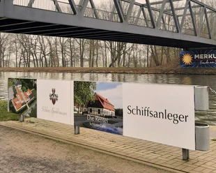 Werbeschild am Wasser unter einer Stahlbrücke; Hinweis auf Schloss, Café und Schiffsanleger.