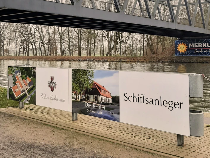 Schiffsanleger Benkhausen Werbeschild am Wasser unter einer Stahlbrücke; Hinweis auf Schloss, Café und Schiffsanleger.