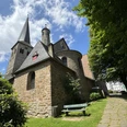 Kirche St. Walburga Steinige Kirche mit spitzem Turm vor blauem Himmel, von Bäumen und Rasen umgeben, grüne Bank davor.