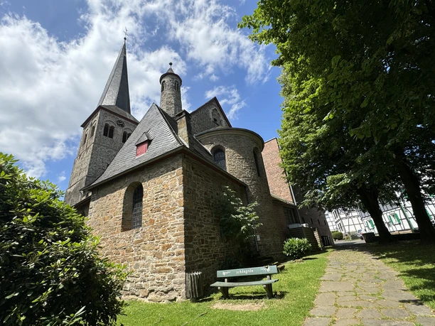 Kirche St. Walburga Steinige Kirche mit spitzem Turm vor blauem Himmel, von Bäumen und Rasen umgeben, grüne Bank davor.