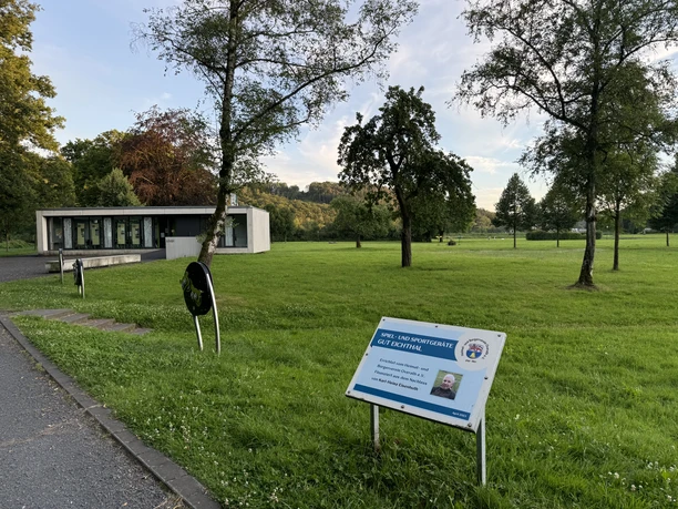 Gut Eichtal Modernes Gebäude im Park, von grüner Wiese und Bäumen umgeben, Infotafel im Vordergrund.