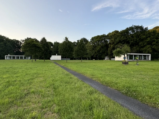 Gut Eichtal Weite grüne Wiese mit zwei modernen, weißen Gebäuden vor einer Baumreihe. Klarer Himmel im Hintergrund.