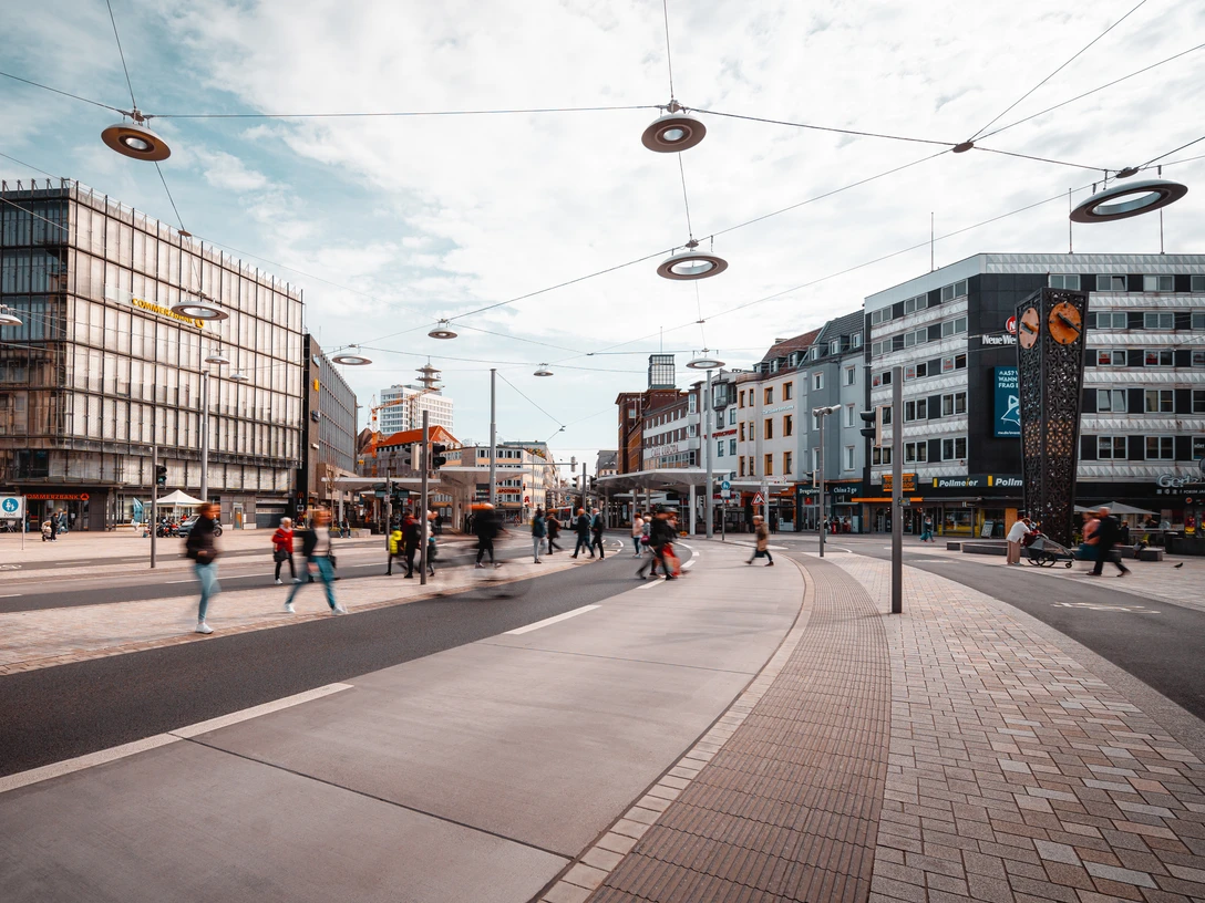 Menschen und Straßenbahnen beleben einen modernen urbanen Platz mit umliegenden Geschäftshäusern.