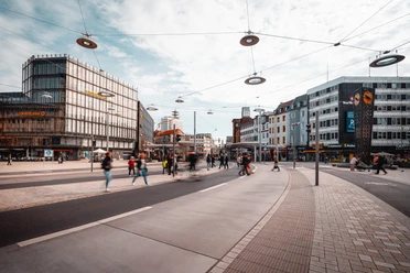 Jahnplatz Bielefeld.jpg Menschen und Straßenbahnen beleben einen modernen urbanen Platz mit umliegenden Geschäftshäusern.