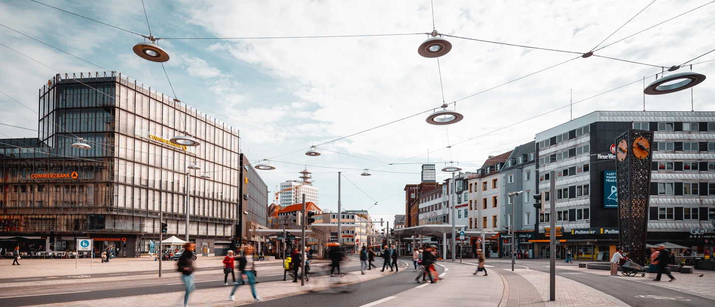 Jahnplatz Bielefeld.jpg Menschen und Straßenbahnen beleben einen modernen urbanen Platz mit umliegenden Geschäftshäusern.