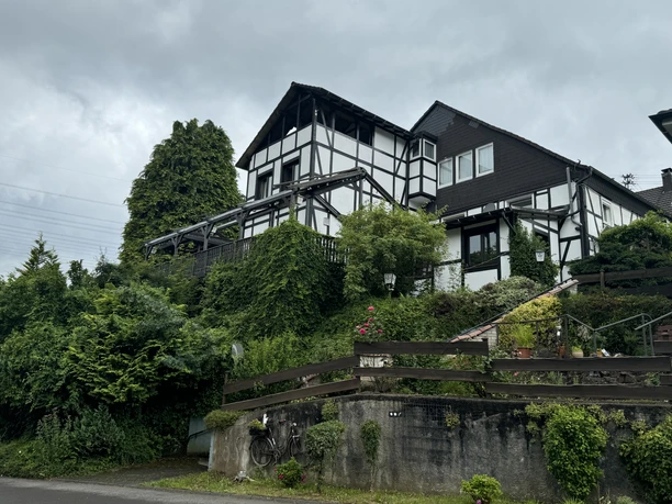 Gasthof Zum Stausee Fachwerkhaus mit üppiger Begrünung an einem Hang, umgeben von Bäumen und bepflanzten Steingärten.