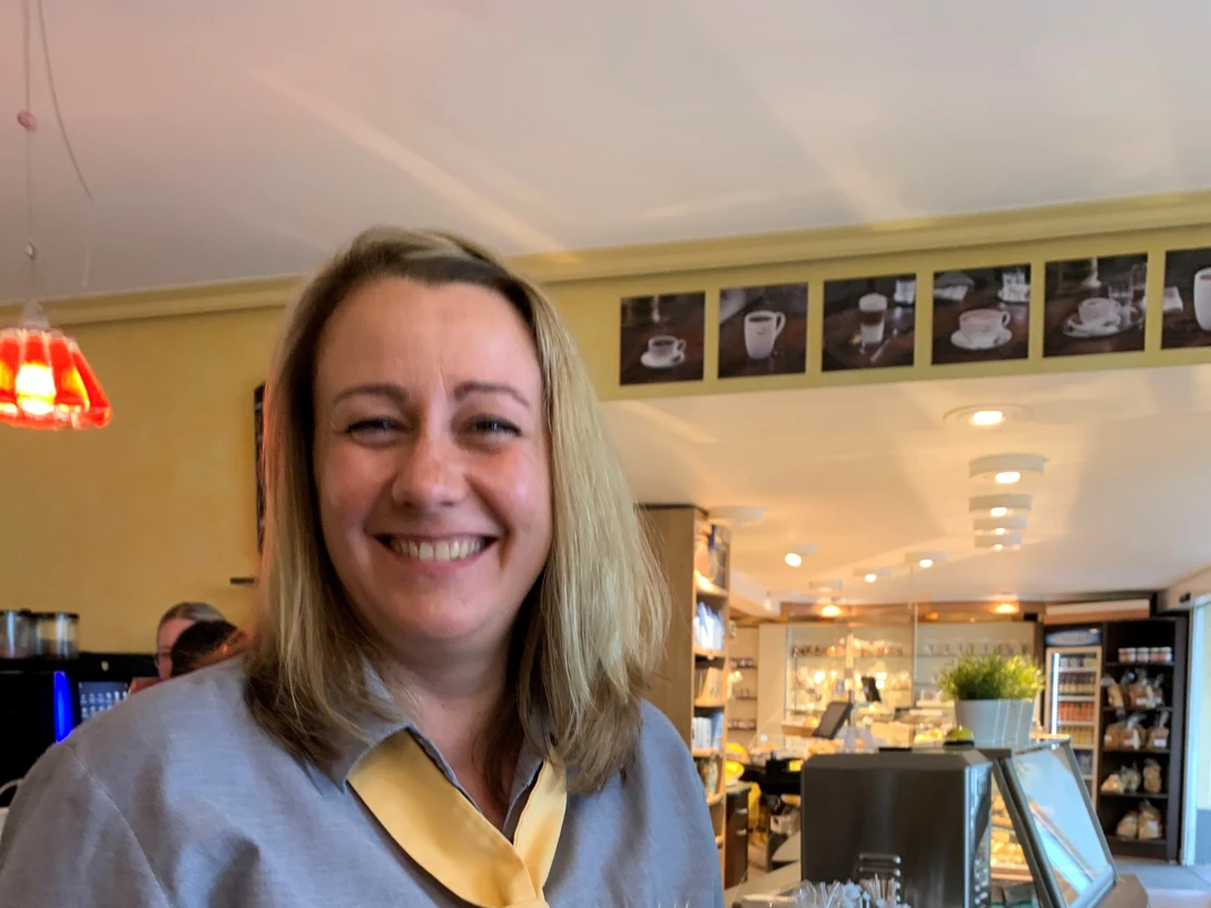 Vorbohle.jpg Eine lächelnde Frau in einem Café hält eine Tüte Gebäck in der Hand.