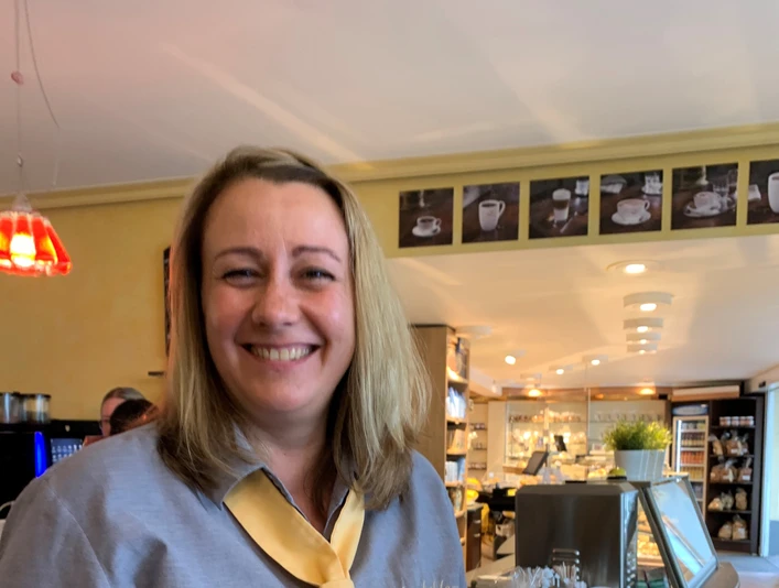 Vorbohle.jpg Eine lächelnde Frau in einem Café hält eine Tüte Gebäck in der Hand.