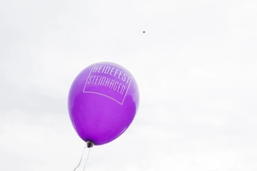 Ein einzelner lila Ballon mit der Aufschrift "Heidefest Steinhagen" schwebt vor wolkigem Himmel.